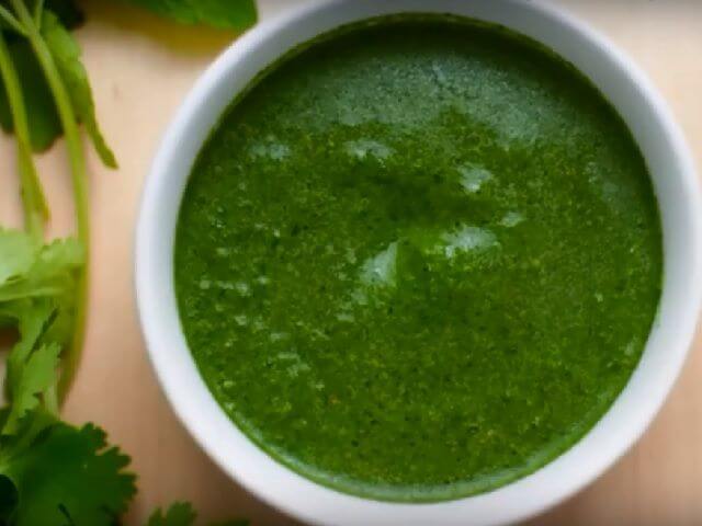 Bowl of vibrant green mint and coriander chutney surrounded by fresh cilantro leaves.
