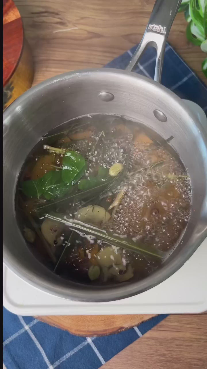 Stahl Artisan Triply Saucepan with Strainer Glass Lid cooking herbs and ingredients in simmering broth.